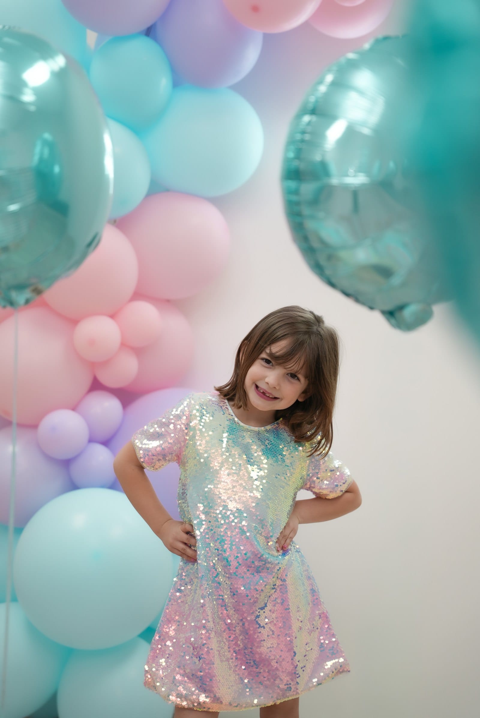 The birthday girl in a sequinned rainbow dress standing proudly in front of a pastel balloon garland — children's birthday photography by Veil & Light in north Jersey