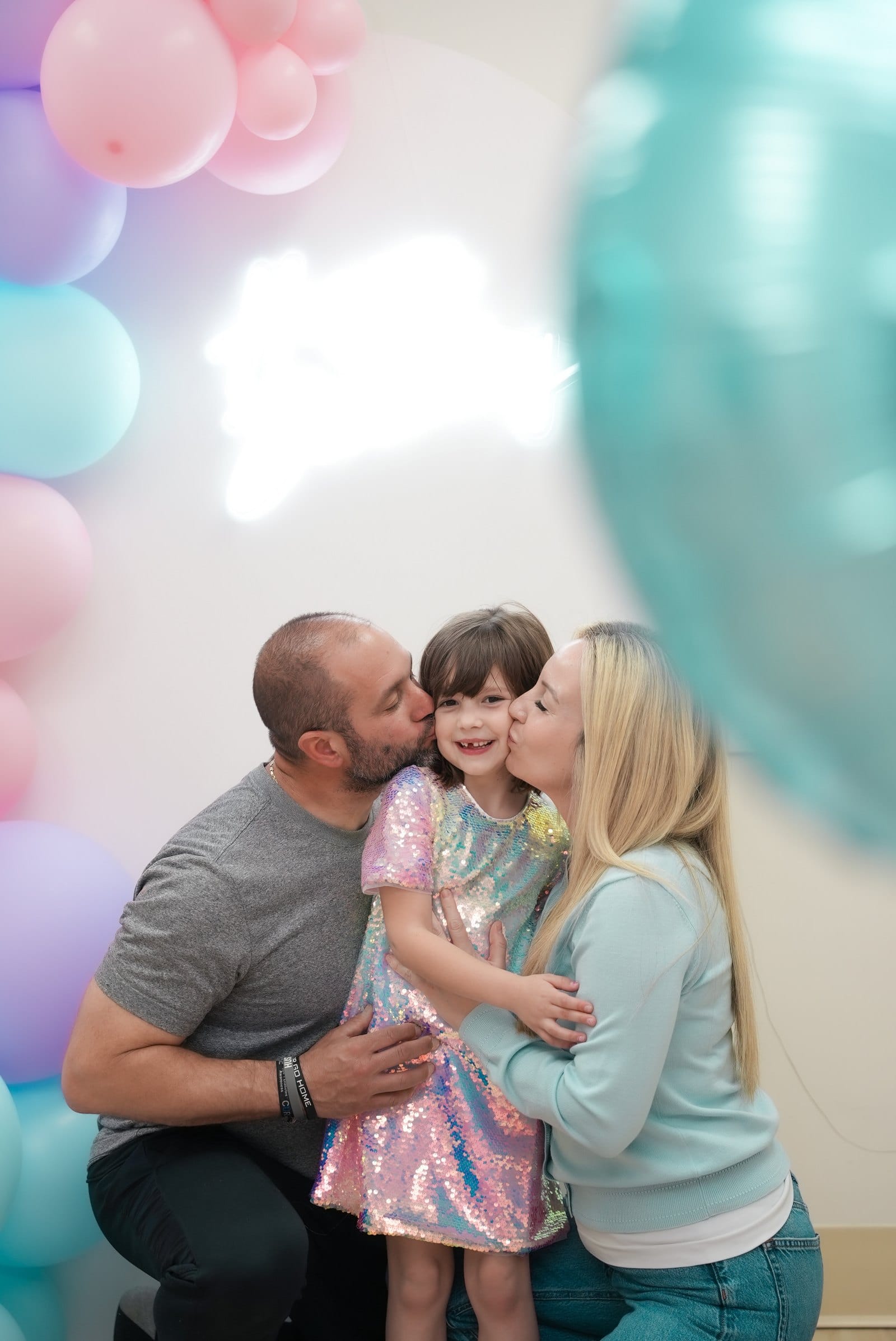 A mother and father each kissing their daughter on the cheek at her birthday party, framed by pink and teal balloons