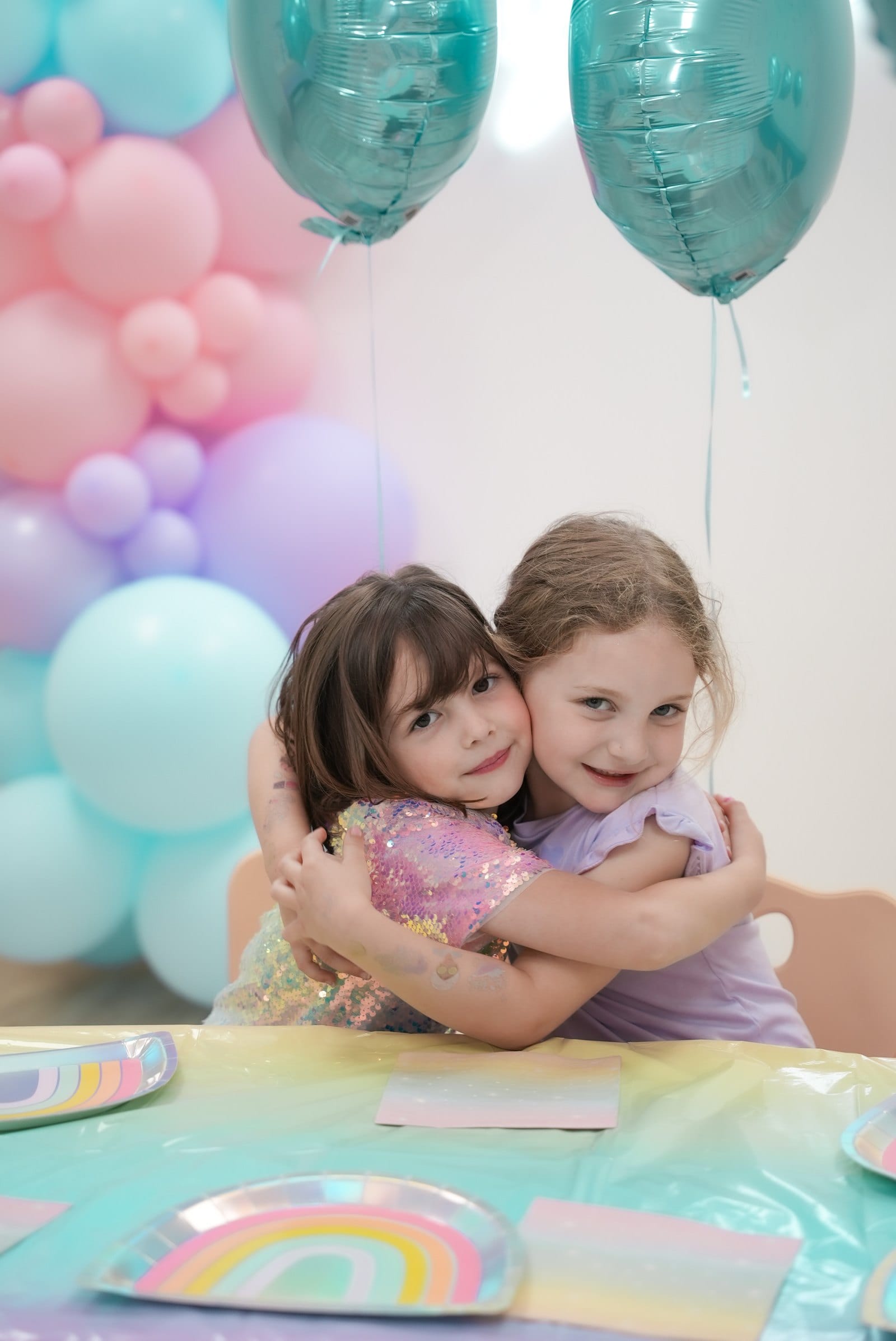 Two young friends hugging tightly at a birthday table, both grinning at the camera in front of a pastel balloon garland