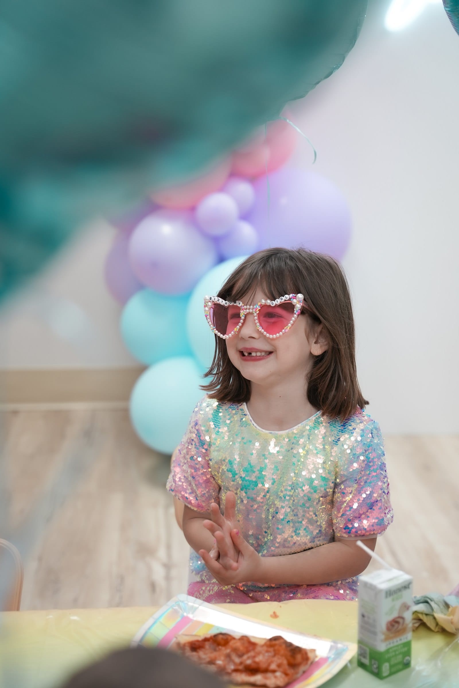 A little girl in heart-shaped sunglasses and a sequinned dress laughing at her birthday table