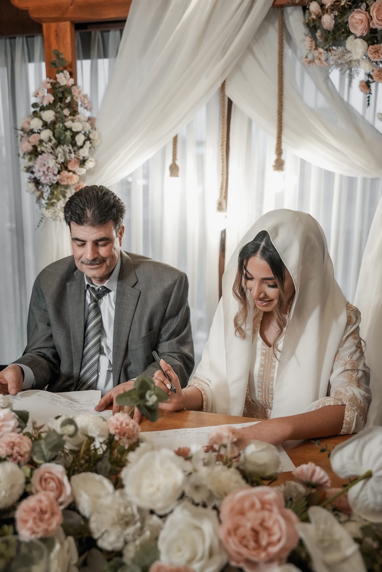 Bride signing the nikkah marriage contract beneath draped white fabric and pink floral arrangements, surrounded by roses — Islamic wedding ceremony photography