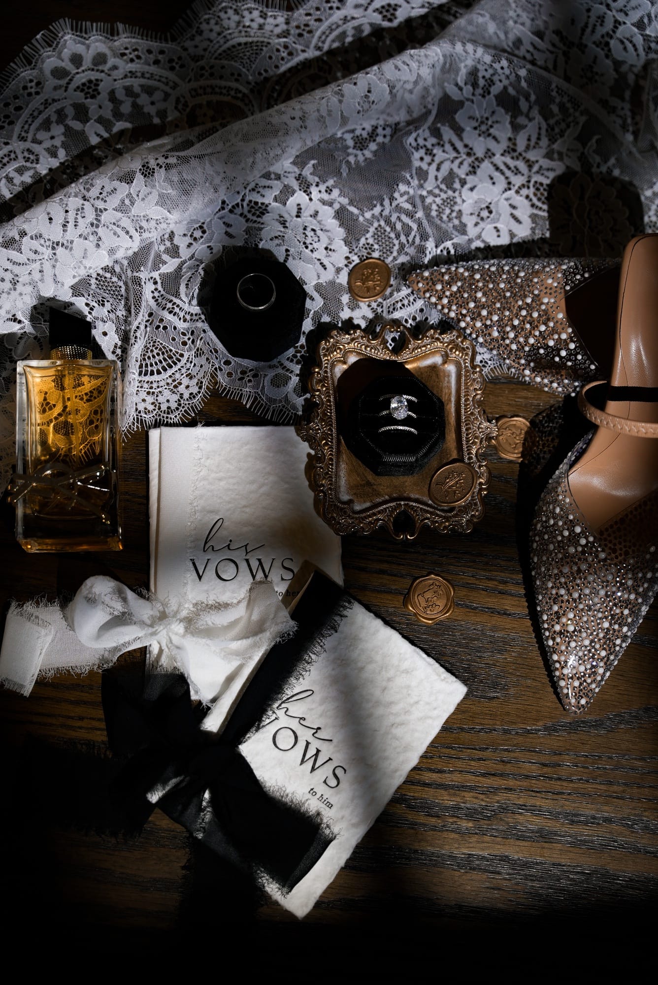 Luxury wedding flat lay — vow books, diamond engagement ring, pearl-encrusted heels, lace, and perfume bottle styled on dark wood