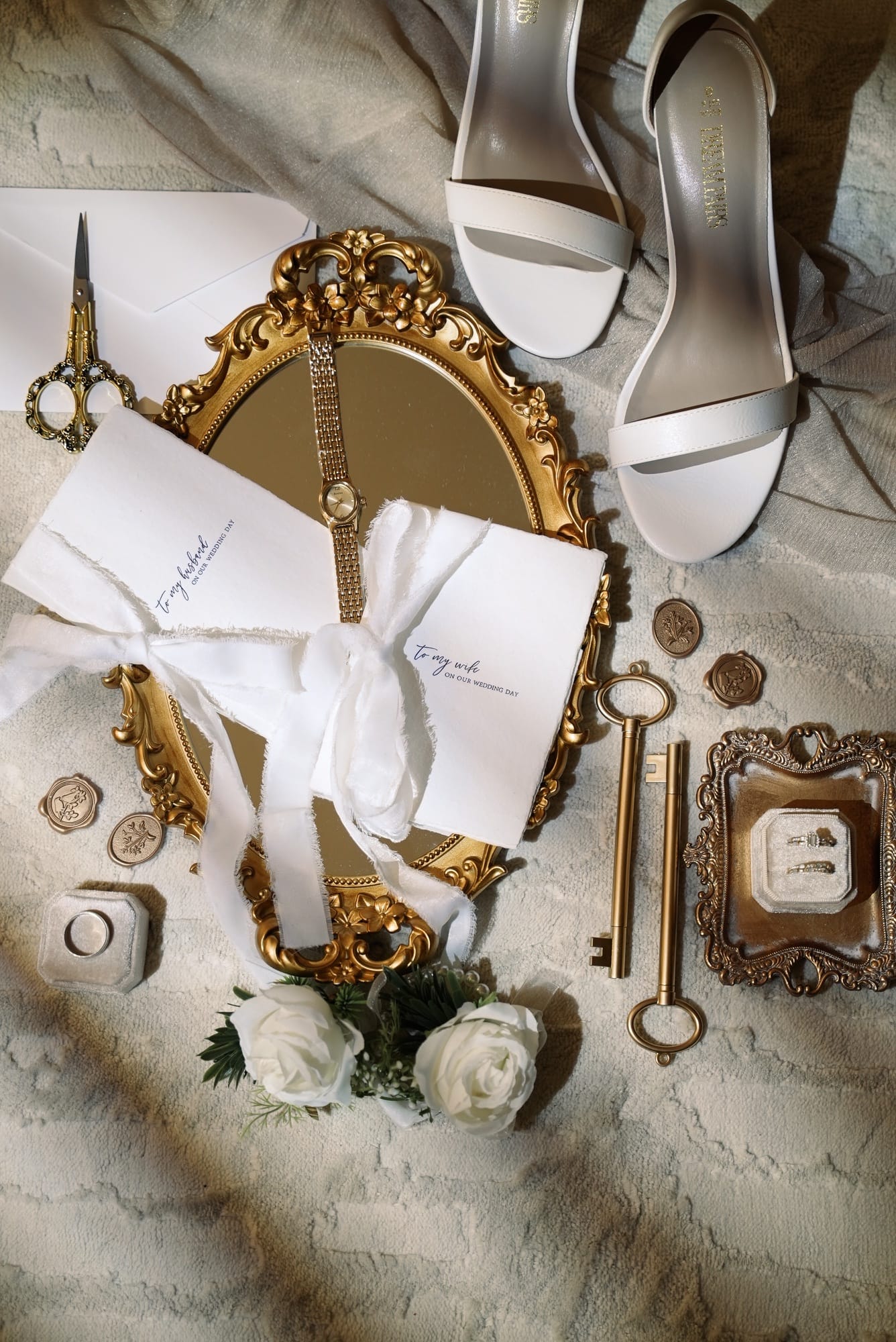 Airy bridal flat lay — vow letters tied with white ribbon, bridal heels, gold vintage mirror, and wedding rings on a velvet tray
