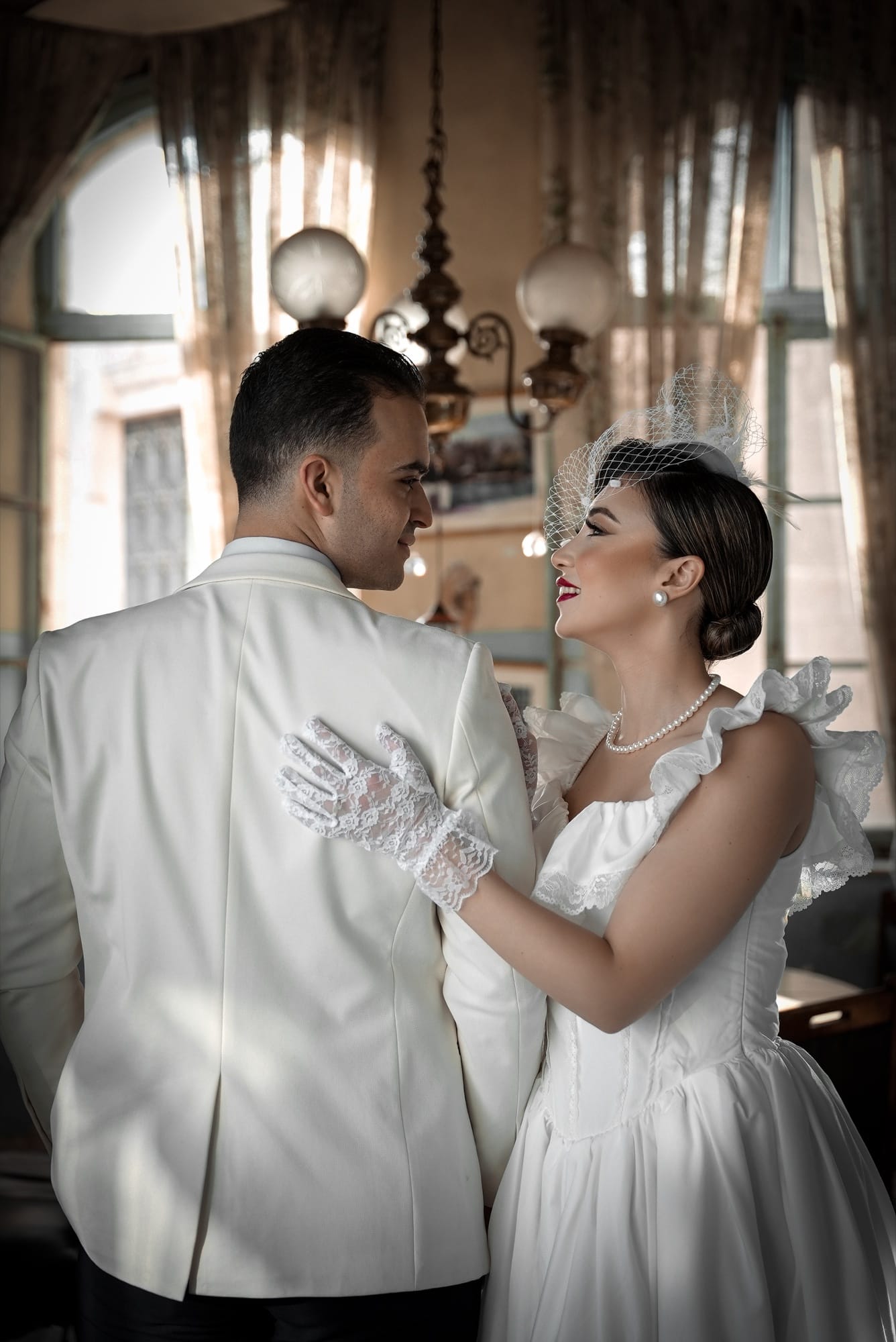 Vintage-styled bride in a retro white dress with lace gloves and birdcage veil, laughing with her groom in a white jacket inside an elegant cafe