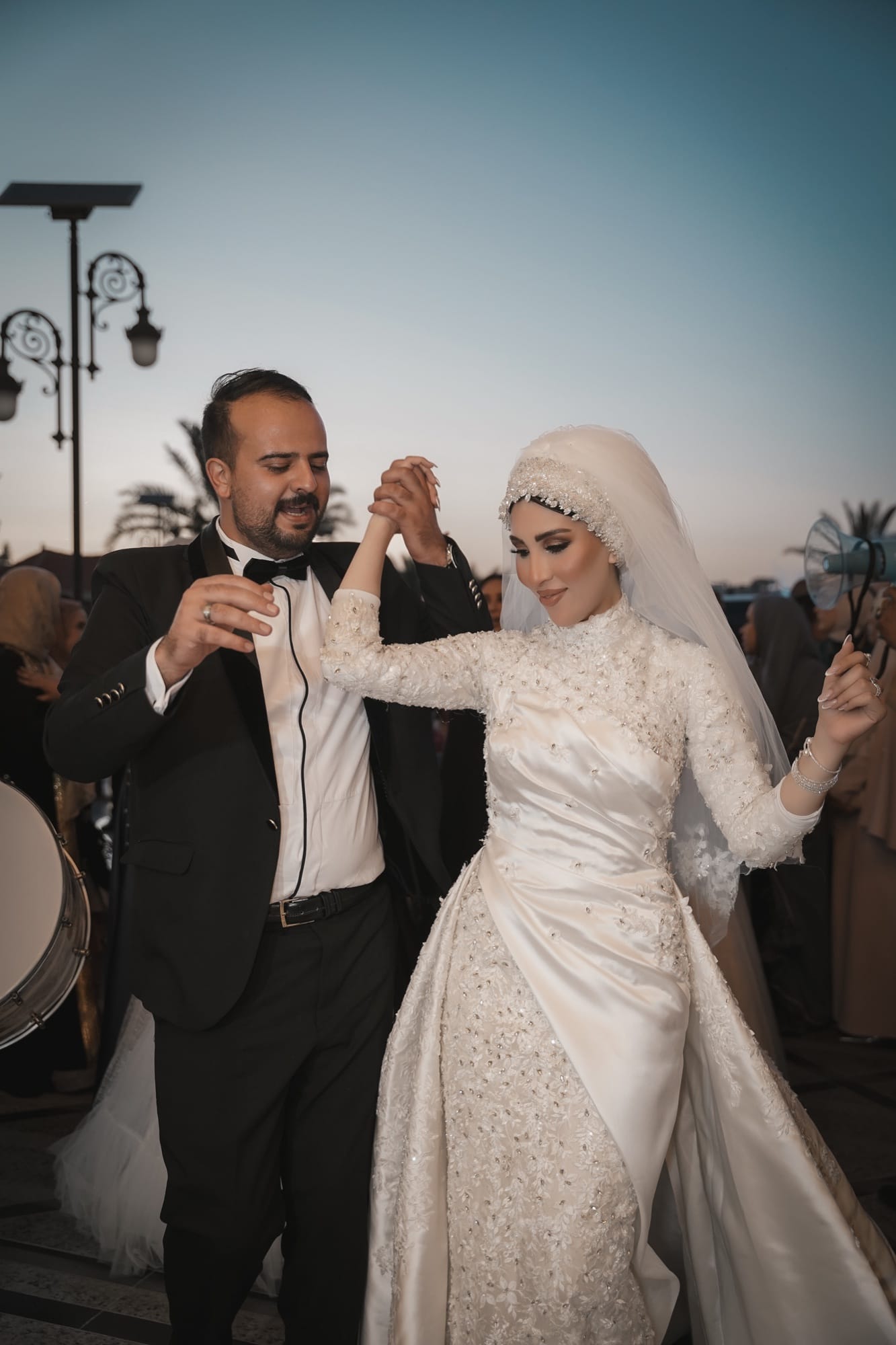 Hijabi bride in a beaded long-sleeve modest gown dancing joyfully with her groom at an outdoor evening reception at dusk