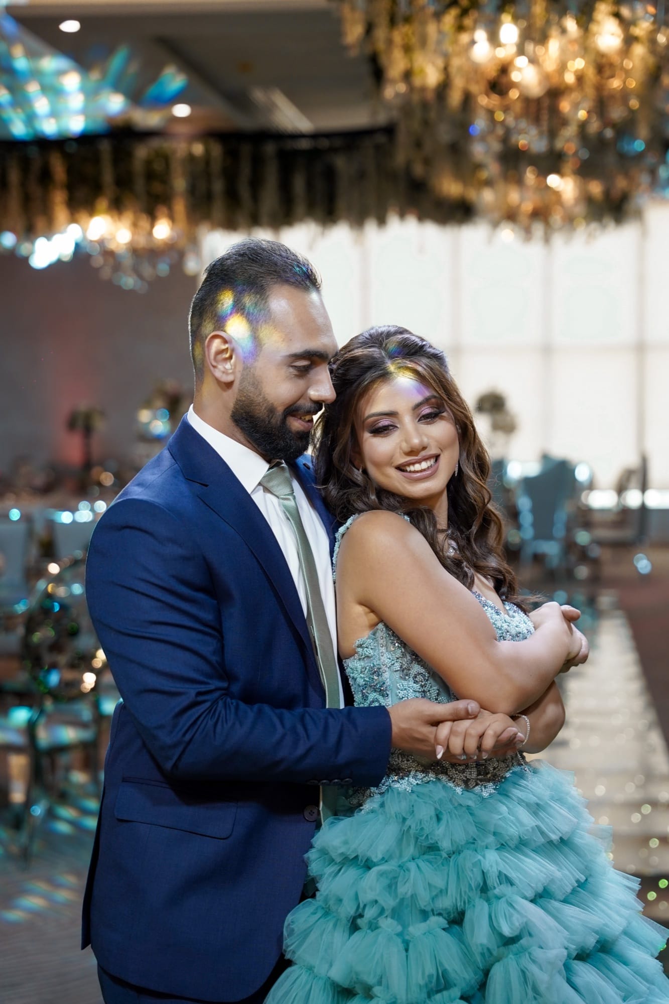Couple embracing and laughing in a grand ballroom, bride in a teal ruffled gown beneath gold crystal chandeliers