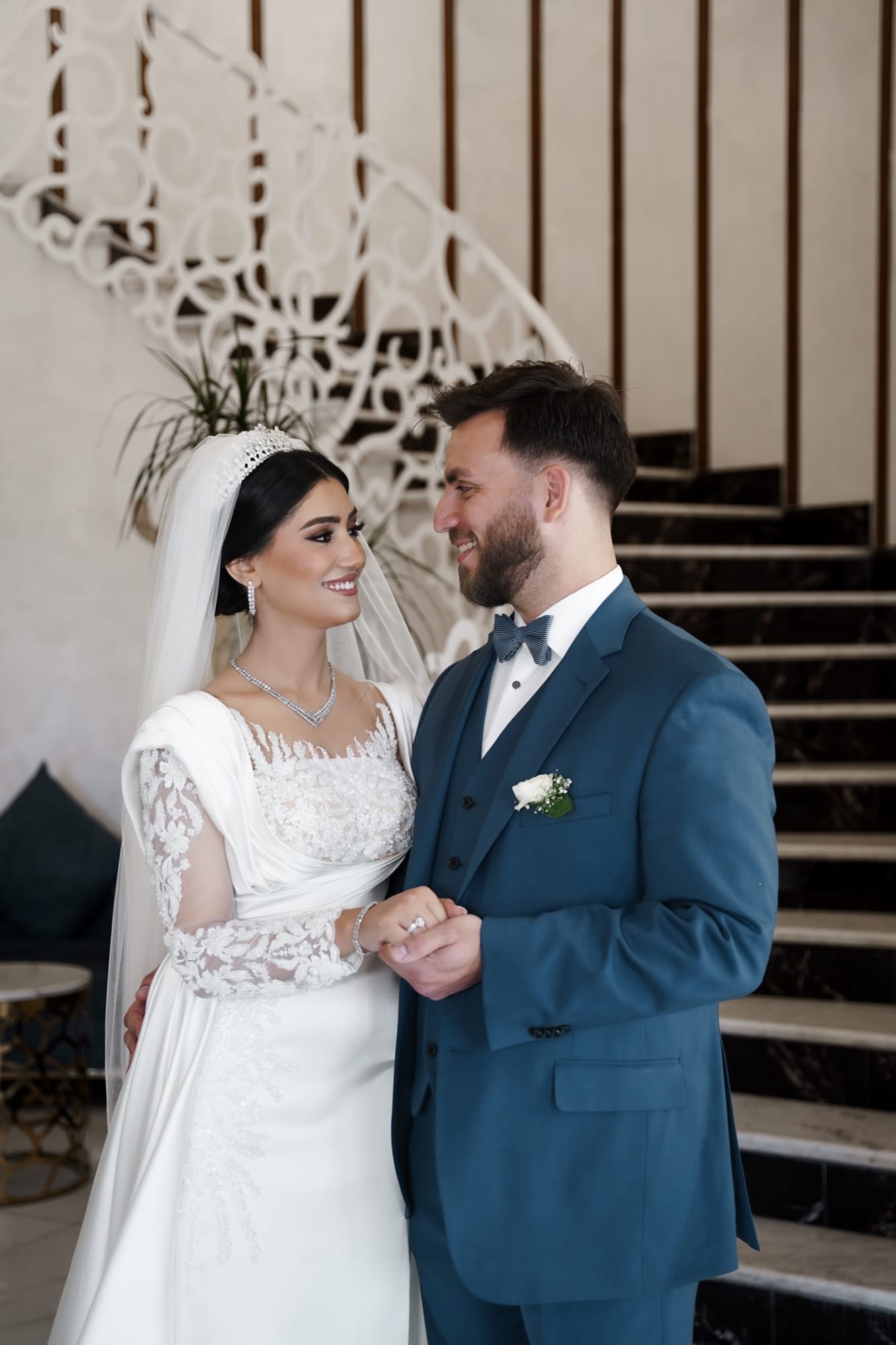 Bride in an elegant long-sleeve lace gown and groom in a steel-blue suit smiling at each other in a luxury venue with an ornate screen backdrop