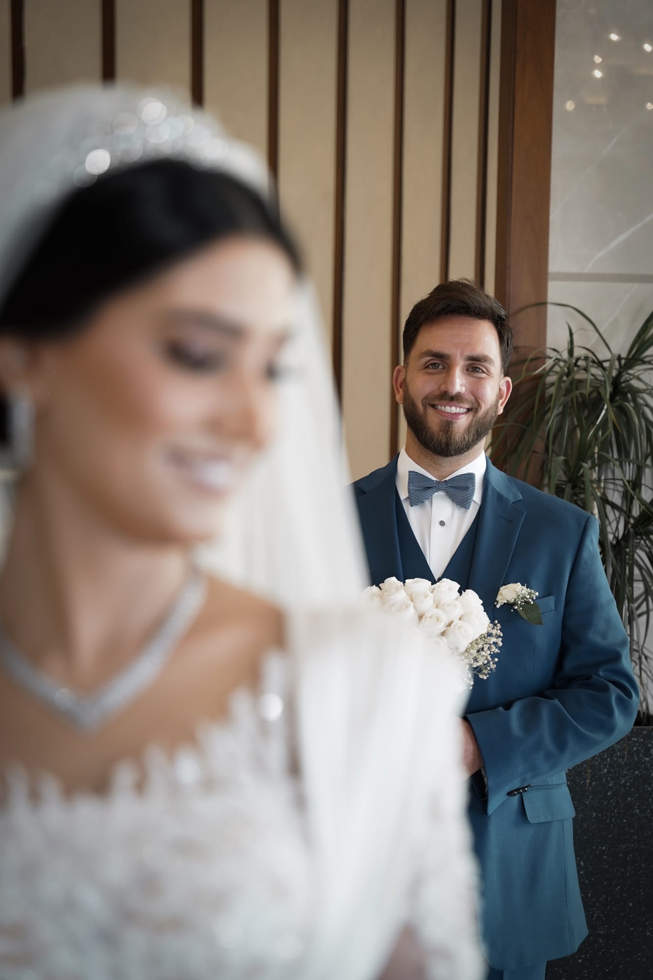 Editorial portrait of the bride in soft focus foreground with the groom smiling behind her, holding a white rose bouquet