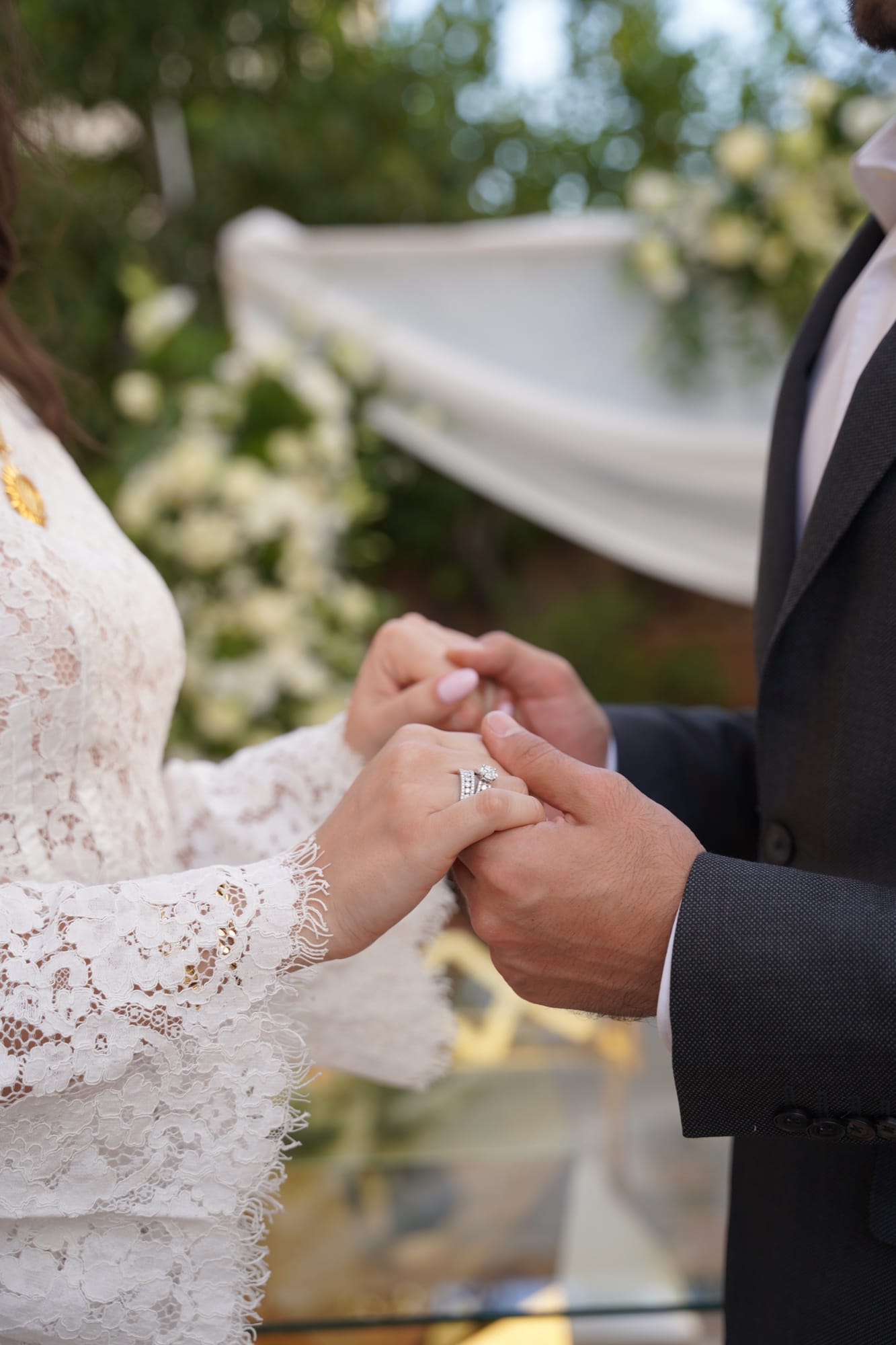 Wedding ring exchange — the bride's lace sleeve and engagement ring against a floral ceremony backdrop