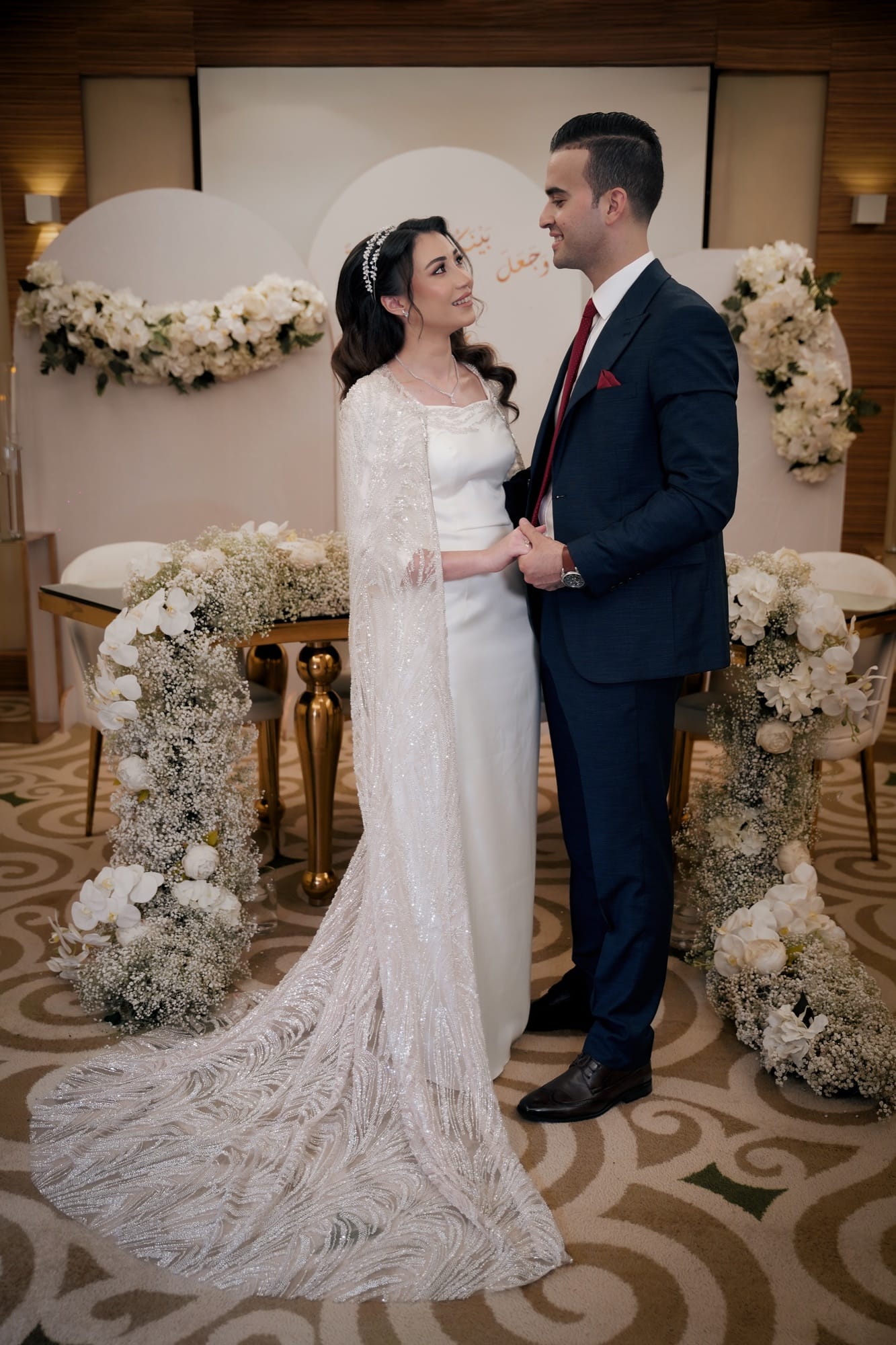 Couple at their nikkah ceremony — bride in a sparkly silver cape gown with a white floral backdrop and baby's breath arrangements