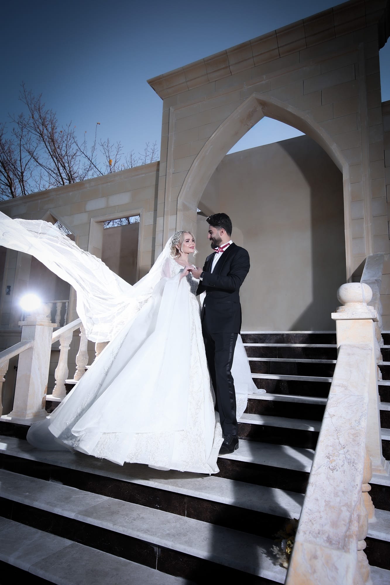 Bride and groom on a grand stone staircase beneath an arched doorway, the bride's cathedral veil billowing dramatically in the night air