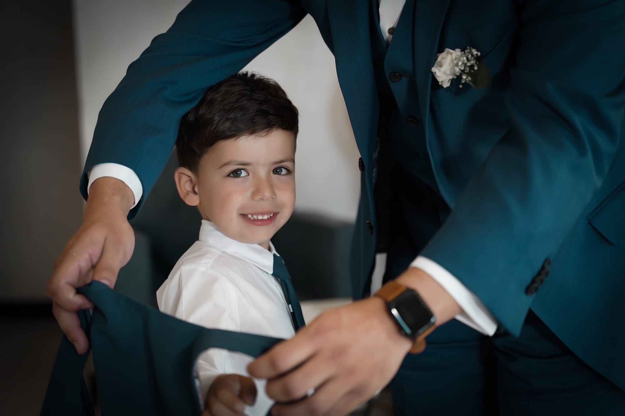A young ring bearer in a teal suit smiling as the groom adjusts his vest before the ceremony