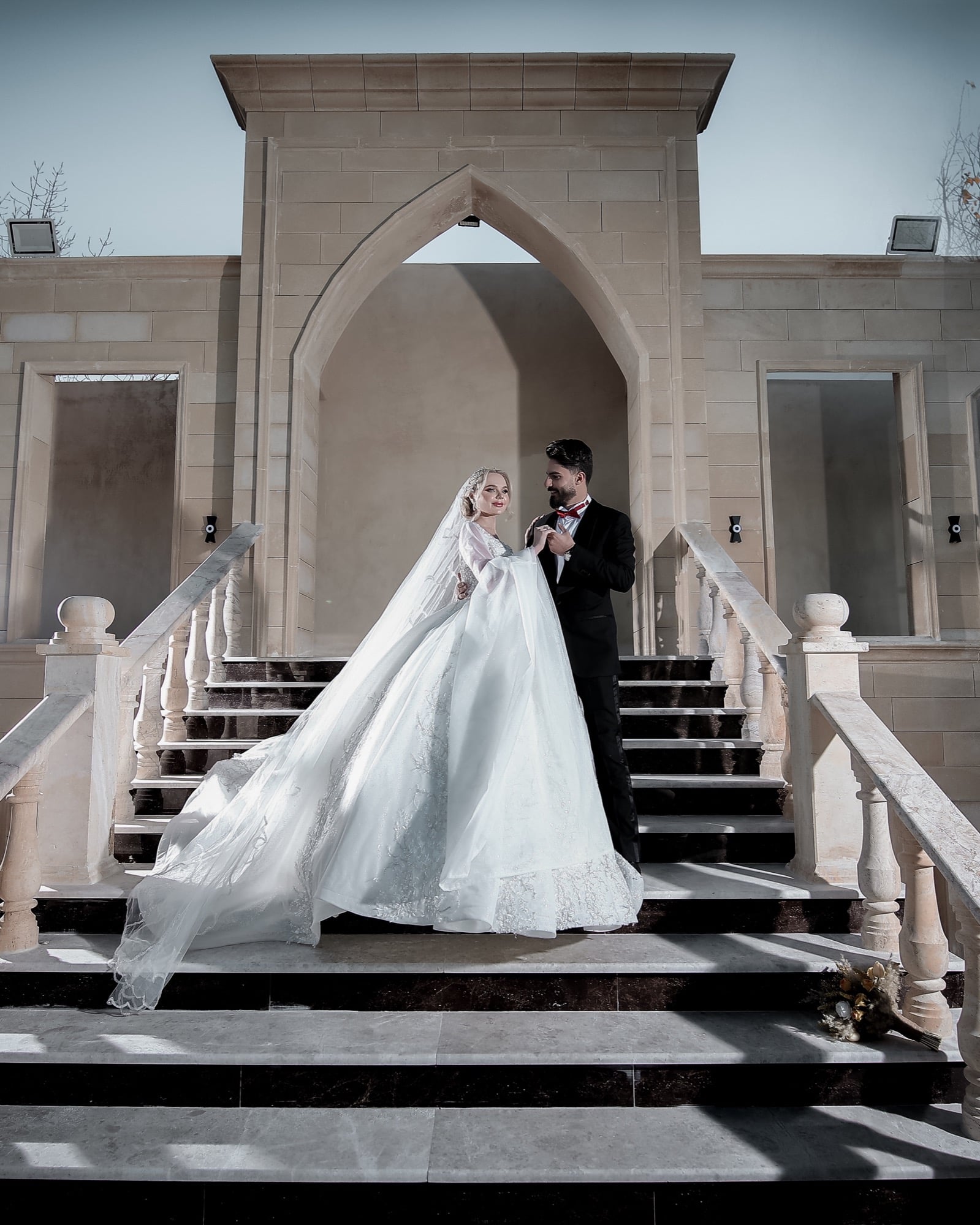 Wedding portrait by Veil & Light — bride and groom paused together on a grand staircase, framed by an arched stone facade and balustrades